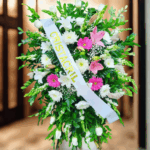 Arreglo floral fúnebre grande con flores blancas, rosas y fucsias, follaje verde abundante y cinta blanca diagonal con texto dorado
