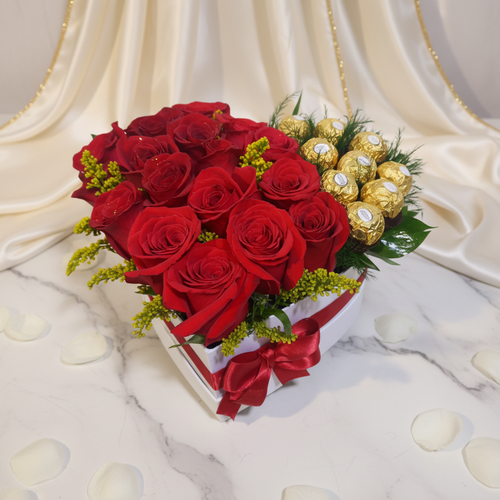 Arreglo en caja de corazón con rosas rojas y chocolates Caja en forma de corazón con rosas rojas frescas y chocolates dorados sobre mesa de mármol con pétalos blancos