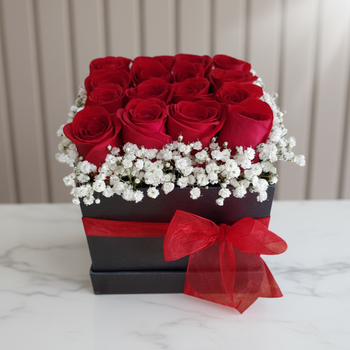 Caja negra con rosas rojas y flores blancas decorativas Arreglo floral de rosas rojas en una caja negra cuadrada con lazo rojo y flores blancas alrededor, sobre mesa de mármol