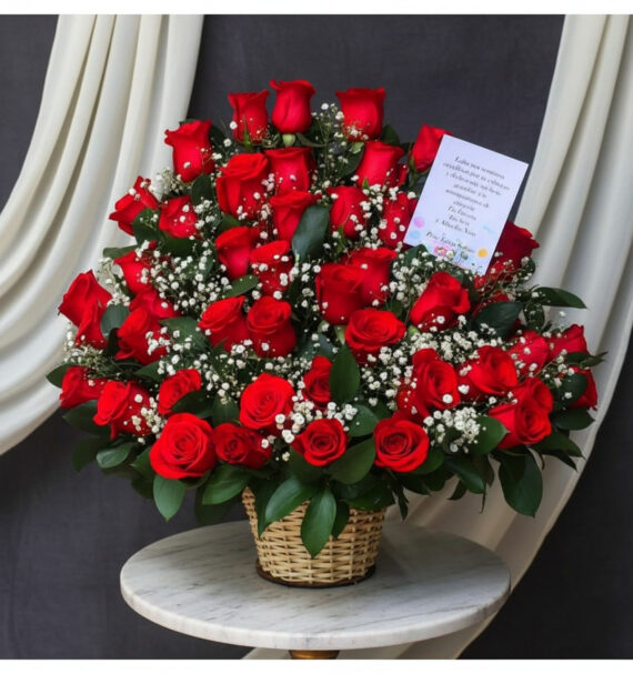 Arreglo de rosas rojas en canasta de mimbre Canasta de mimbre sobre mesa redonda con gran arreglo de rosas rojas y flores blancas de relleno, acompañadas de tarjeta sobre fondo de cortinas blancas y tela gris