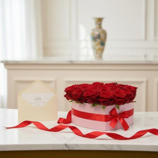 Arreglo floral CAMILA con 24 rosas rojas premium en caja de corazón rosa, cinta roja y tarjeta de regalo elegante sobre mármo