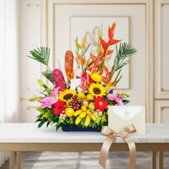 Arreglo floral exótico con heliconias, girasoles, lirios y frutas tropicales, acompañado de una tarjeta de regalo y cinta sat