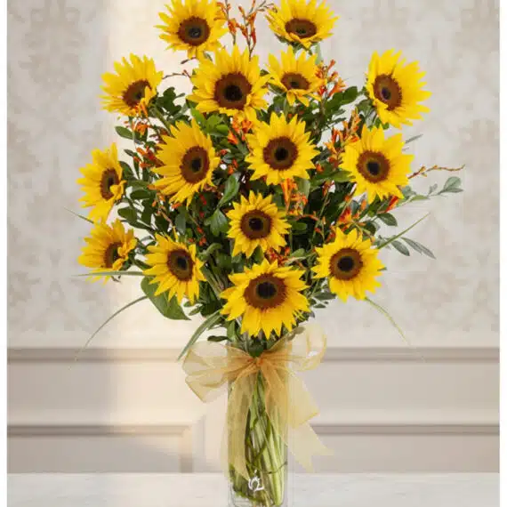 Arreglo floral de girasoles LENU en un jarrón de cristal con lazo dorado, sobre una mesa de mármol y fondo elegante.