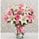 Arreglo floral vibrante con rosas rosadas, lirios blancos y claveles rosados en un jarrón de cristal con lazo rosa, ideal par