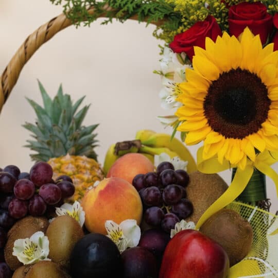 Primer plano de la Canasta de Frutas Draco, destacando un girasol vibrante, rosas rojas, uvas, kiwis, duraznos y piñas, con g